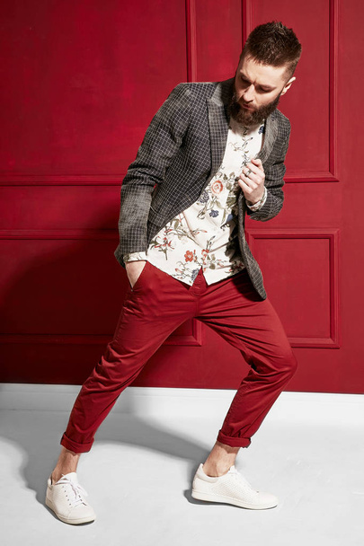 Fashion portrait of handsome man with dark hair and beard wearing red pants. floral shirt and plaid jacket. Red background - Valokuva, kuva