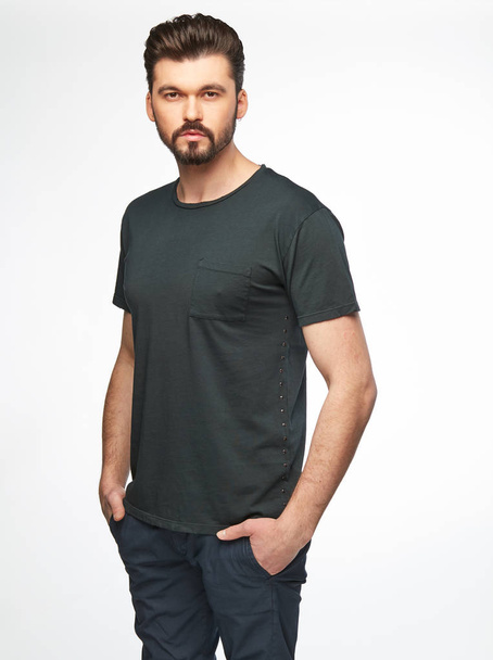 Fashion portrait of handsome male model with dark hair, beard and eyes, wearing grey t-shirt and pants and posing on white background - Fotoğraf, Görsel