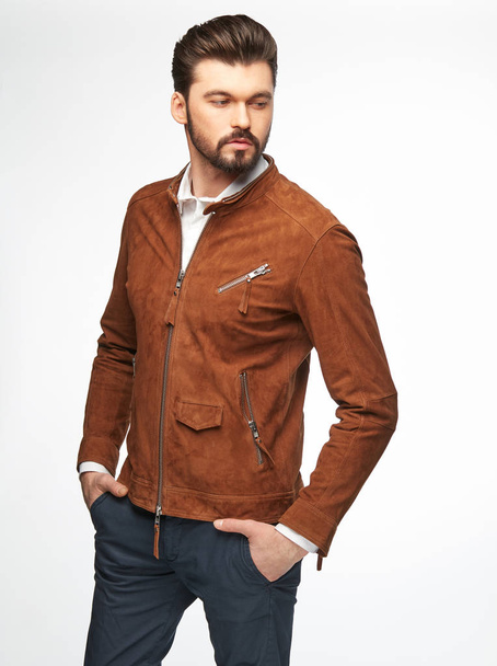 Fashion portrait of male model with dark hair, beard and eyes, wearing white shirt, orange brown jacket and dark pants, posing on white background - Foto, imagen