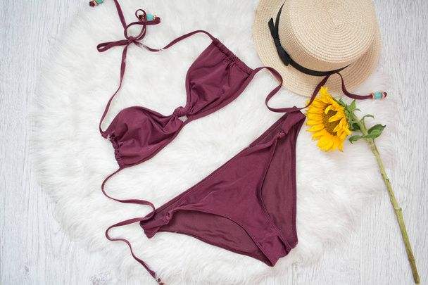 Burgundy swimsuit, straw hat and sunflower. White fur on the background, top view - Photo, Image