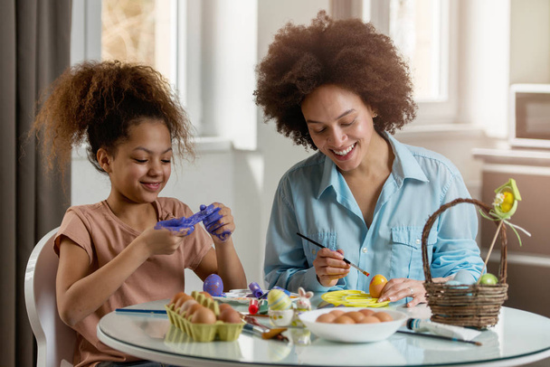 Beautiful African American woman and her daughter coloring Easter eggs at the table   - Foto, afbeelding