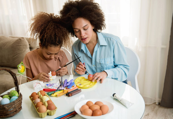 Beautiful African American woman and her daughter coloring Easter eggs at the table   - Фото, зображення