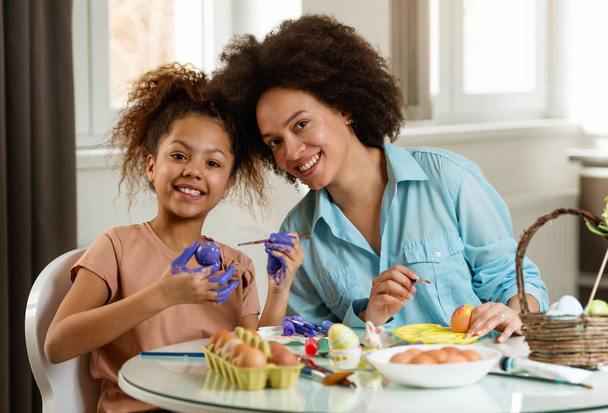 Beautiful African American woman and her daughter coloring Easter eggs at the table   - Photo, Image