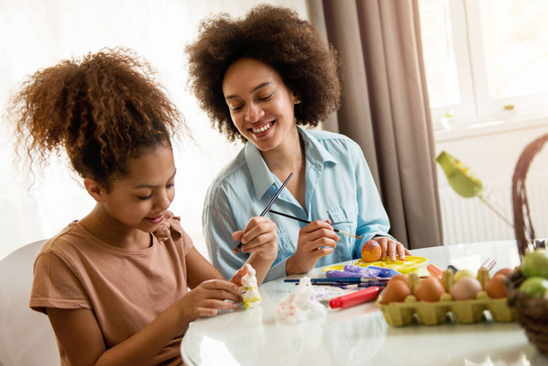 Beautiful African American woman and her daughter coloring Easter eggs at the table   - Foto, immagini
