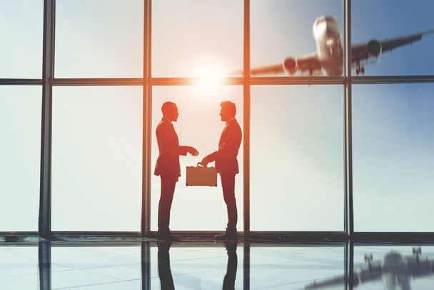 Silhouette view of two young businessmen are standing with suitcase in modern office with panoramic windows - Foto, afbeelding