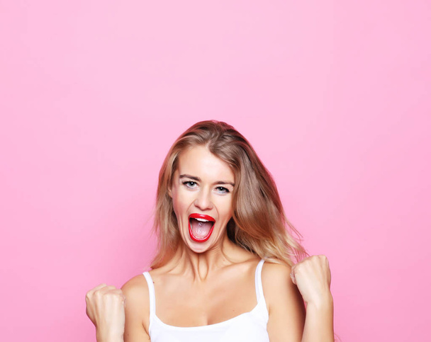 successful young blond woman with raised hands shouting and celebrating success over pink background - Photo, Image