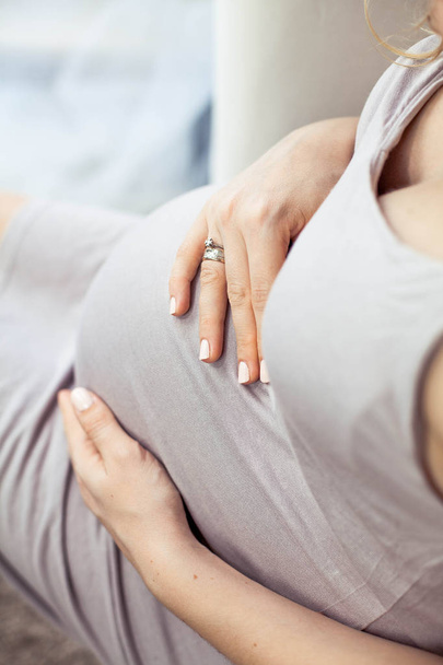 eine junge schöne schwangere Frau sitzt auf einem Sofa und berührt ihren Bauch. Schwanger in einem schönen Kleid. Mutterschaft. Schwangerschaft. Porträt einer glücklichen Schwangeren. gemütlich. - Foto, Bild