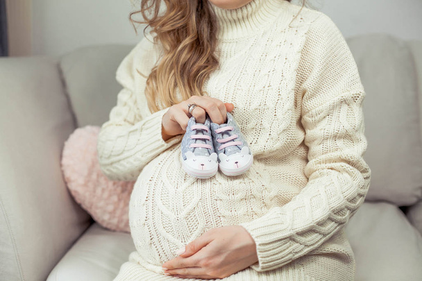 A young beautiful pregnant woman touches her tummy. A pregnant woman in a knitted dress sits on the sofa and holds children's shoes. Motherhood. Pregnancy. Portrait of a happy pregnant woman. Cozy. - Foto, Imagem