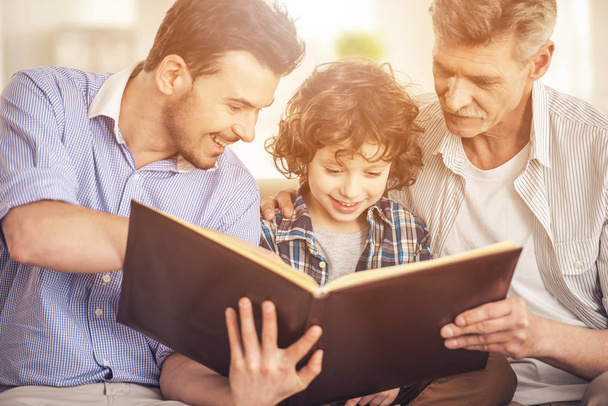 Generation portrait. Grandfather, father and son sitting and reading a book on sofa - Photo, image