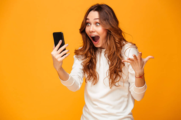 Shocked brunette woman in sweater holding smartphone and rejoices - Фото, изображение