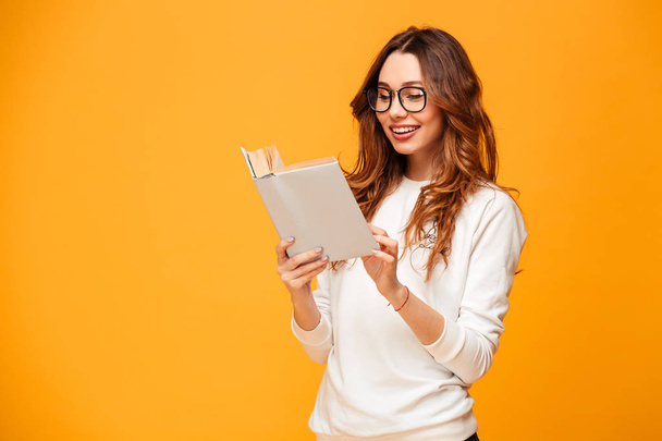 Smiling brunette woman in sweater and eyeglasses reading book - Fotó, kép