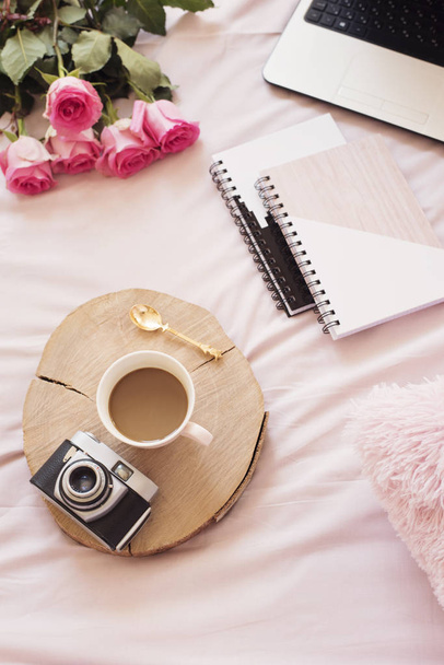 Coffee, old vintage camera in bed on pink sheets. Roses, notebooks and laptop around. Freelance fashion home femininity workspace in flat lay style - 写真・画像