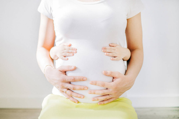 Giovane donna incinta in t-shirt bianca e mani di bambino che toccano il suo stomaco. Concetto famiglia felice
. - Foto, immagini