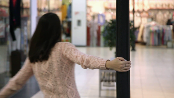 Cheery girl turns around a metallic pole and dances in a glassy shopping mall                            A hilarious view of a stylish young woman with sportive figure and long loose hair who turns around a pole and dances in a funny way in a malll - Footage, Video
