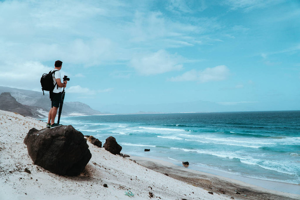 Traveler with camera staying on the black volcanic boulder and admiring unusual barren landscape of Baia Das Gatas beach. North of Calhau, Sao Vicente Island Cape Verde - 写真・画像