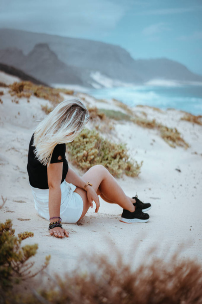 Young woman sitting on white sand dune surrounded by barren desert vegetation. Atlantic ocean coastline in background. Sao Vicente Cape Verde - Foto, immagini