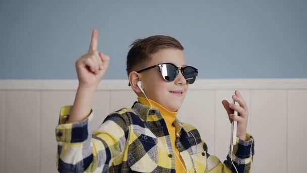 A handsome guy in sunglasses listens to music from the phone in white headphones and he fun dancing in the white background at home. Indoors. Technology - Кадри, відео