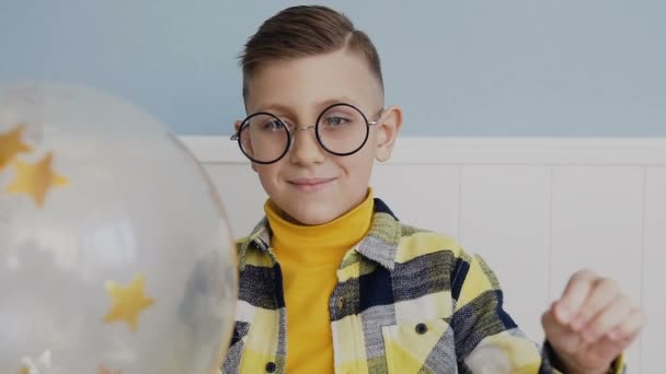 Portrait. A handsome boy in round glasses holds the balloon and pierces it with a needle. Inside the balloon is a golden stars. In the room, on a white background - Materiaali, video