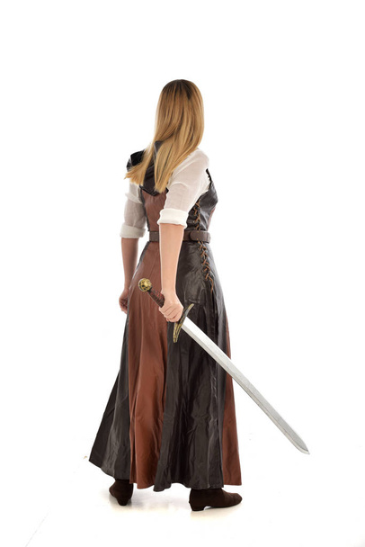 retrato de comprimento total da menina vestindo traje de fantasia marrom, segurando uma espada longa, em pé posar no fundo do estúdio branco
.  - Foto, Imagem