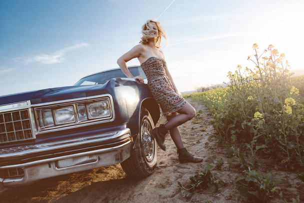 Road trip. Young woman standing by a V8 American car at sunset. - 写真・画像