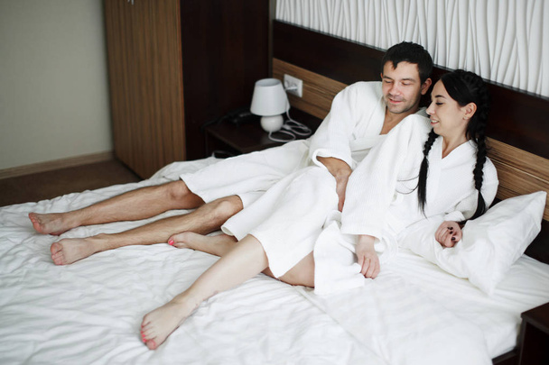 Jovem marido e mulher de casacos brancos brincar e sorrir na cama em um
 - Foto, Imagem