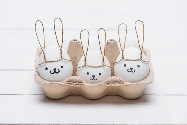 пасхальные куриные яйца с улыбками и кроличьи уши в лотке для яиц на столе
 - Фото, изображение