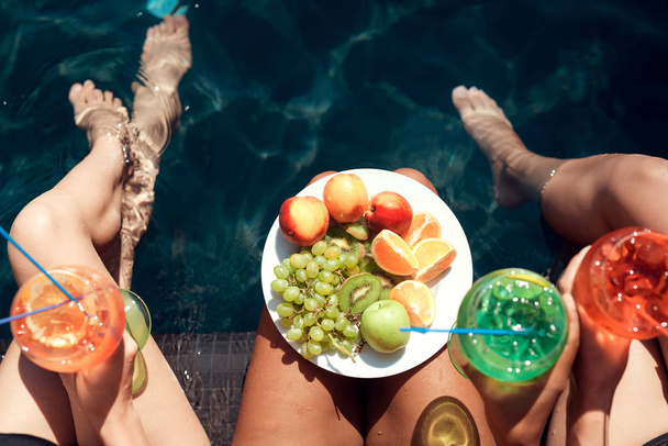Bovenaanzicht. Meisjes in zwempakken eten fruit en drinken cocktails in het zwembad in de zomer. Zomervakantie vrienden samen bij zwembad feest. - Foto, afbeelding