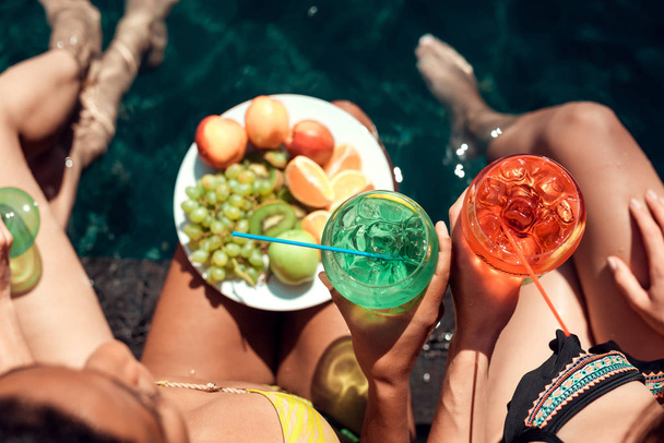 Top view. Girls in swimsuits eat fruits and drink cocktails in swimming pool in summertime. Summer vacation friends together at swimming pool party. - Fotoğraf, Görsel