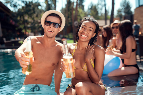 Young man is sunglasses and cheerful woman cheering with beer bottles in swimming pool. Company of young people spend weekend in pool. - Valokuva, kuva
