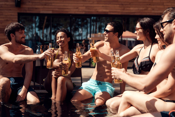 Company of friends cheering with beer bottles in swimming pool. Swimming pool party. Company of young people spend weekend in pool. - Foto, afbeelding