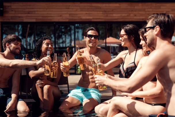 Company of friends cheering with beer bottles in swimming pool. Swimming pool party. Company of young people spend weekend in pool. - Photo, Image