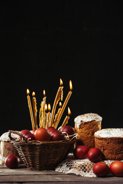окрашенные куриные яйца в корзину, горящие свечи и домашние пасхальные торты на черном
 - Фото, изображение