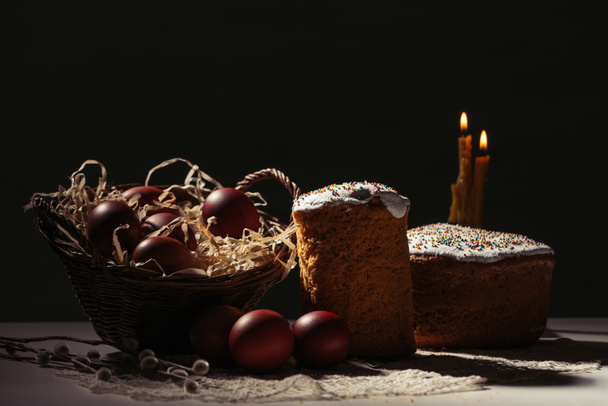basket with painted chicken eggs, easter cakes and burning candles on black - Foto, Bild