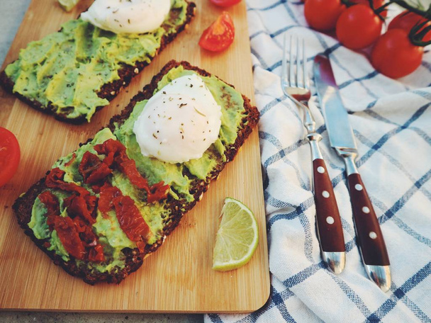 Close view of sandwiches with mashed avocado, poached eggs, lime slices and cherry tomatoes served with knife, fork and napkin - Photo, Image