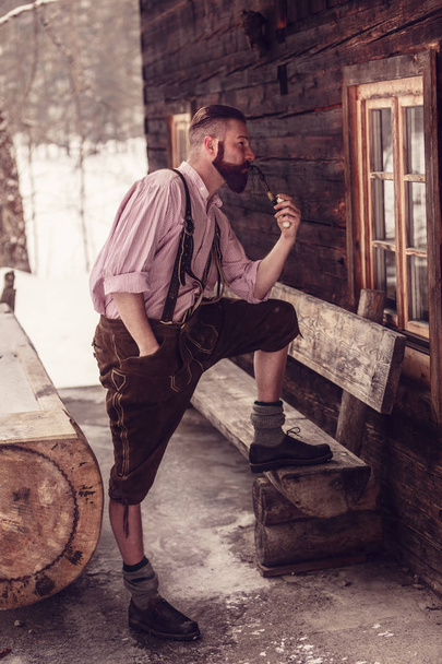 A Bavarian man in lederhosen enjoys his pipe in front of his log cabin in the wilderness - Foto, afbeelding