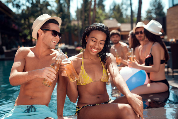 Young man is sunglasses and cheerful woman cheering with beer bottles in swimming pool. Company of young people spend weekend in pool. - Foto, Bild