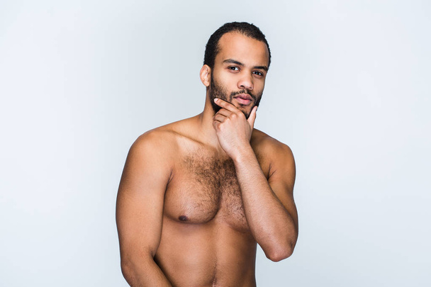 Handsome and confidant. Portrait of handsome shirtless young black man keeping hand on chin and looking at camera while standing against white background - Photo, image
