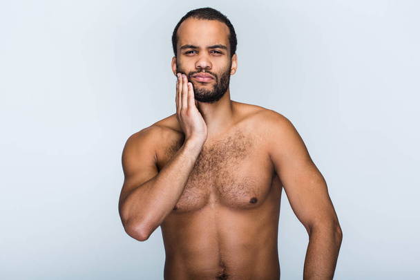Зубная боль. Портрет красивого молодого чернокожего мужчины без рубашки, смотрящего в камеру и трогающего подбородок, стоя на белом фоне
 - Фото, изображение