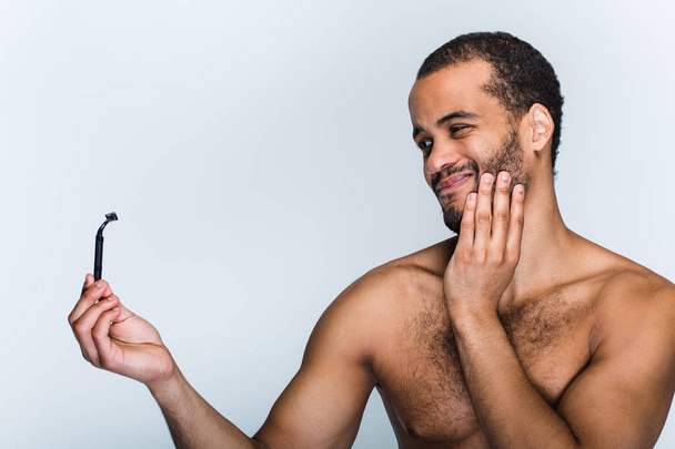 Painful method. Portrait of handsome shirtless young black man looking at razor while standing against white background - Photo, Image
