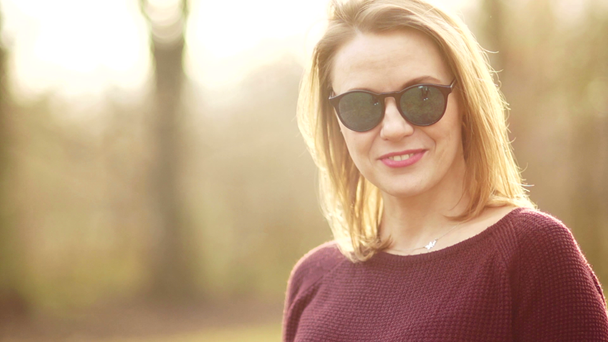 A cute middle-aged woman looks into the camera strictly and benevolently smiling. She wears sunglasses. A girl rests in the city park in early spring. Spring sunset - Footage, Video
