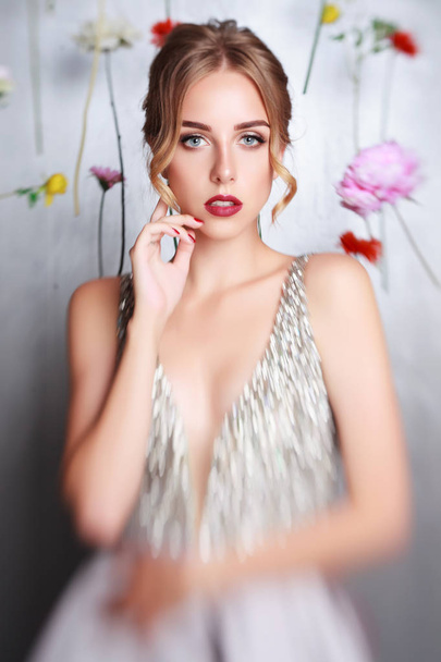 beautiful young girl in luxury silver dress. hanging colourful flowers. blonde with bridal hairstyle. idea for wedding decor. spring portrait for young lady - Foto, Imagem