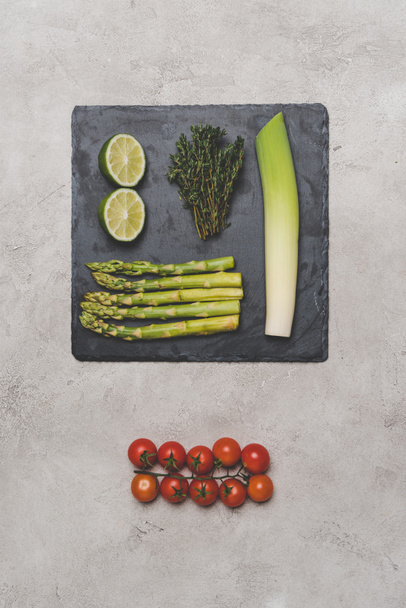 top view of tomatoes, lime, leek and asparagus on slate board on grey - Фото, изображение