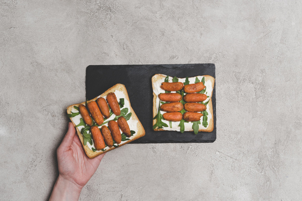 cropped shot of human hand and healthy sandwiches with arugula and baby carrots on slate board - Photo, Image