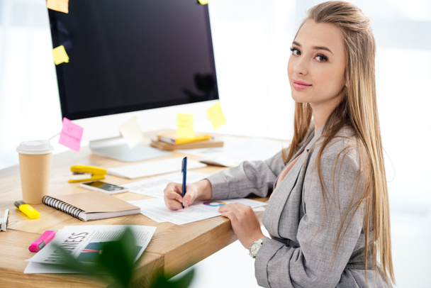 vista lateral de la mujer gerente de marketing mirando a la cámara mientras hace papeleo en el lugar de trabajo en la oficina
 - Foto, Imagen