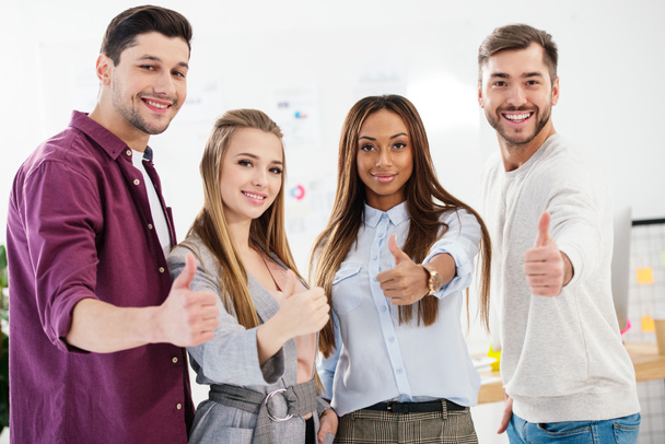 portrait of young smiling multiracial business people showing thumbs up and looking at camera in office - 写真・画像