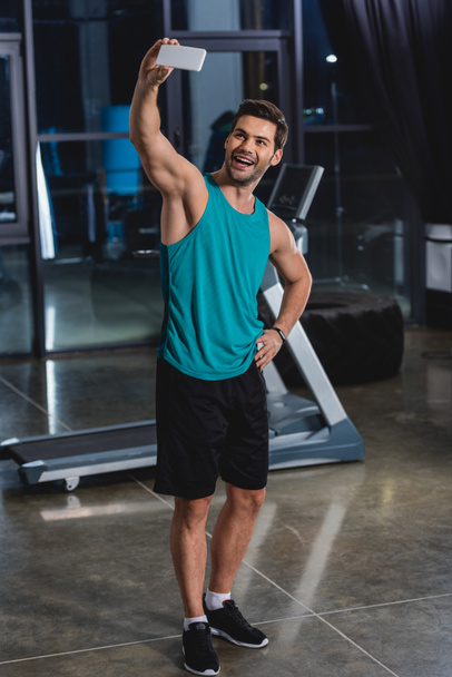 cheerful sportsman taking selfie on smartphone in gym with treadmill - Фото, изображение