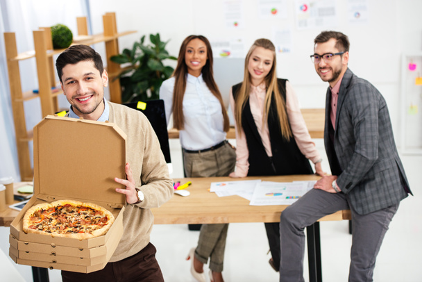 seçici odak pizza el ve Office arkasında çok ırklı meslektaşları ile gülümseyen iş adamı - Fotoğraf, Görsel
