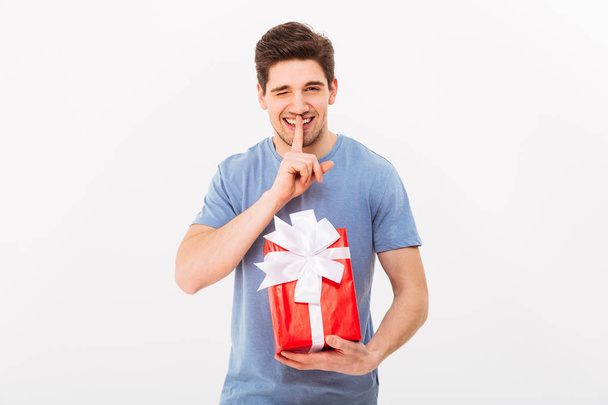 Young man 30s holding gift box with bow and keep secret with fin - Фото, зображення