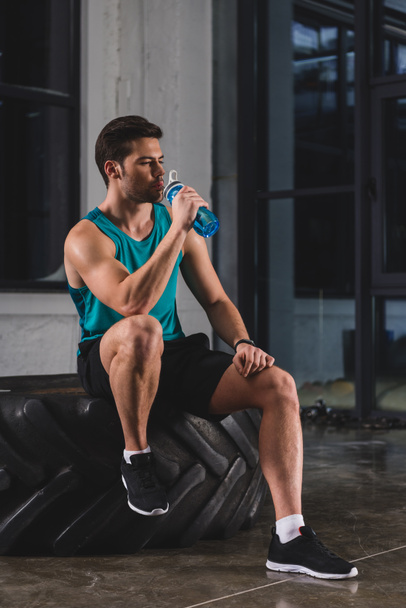 sportsman sitting on tire and drinking water after cross training in gym - Fotoğraf, Görsel