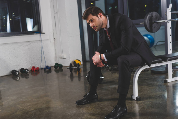 уставший бизнесмен в костюме с фитнес-трекером сидит в тренажерном зале с гантелями
 - Фото, изображение